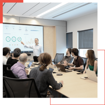 A man is presenting data on a screen to a group of seven people seated around a conference table in a modern office meeting room, showcasing insights from Google Chronicle as part of their managed detection and response strategy.