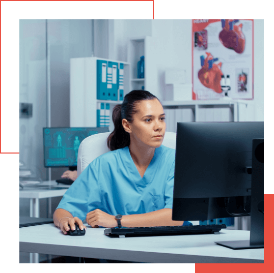  A person in medical scrubs sits at a desk working on a computer, possibly analyzing data with Google Chronicle. A second individual in the background is also engaged with their computer. Medical posters decorate the walls, adding to the professional atmosphere.