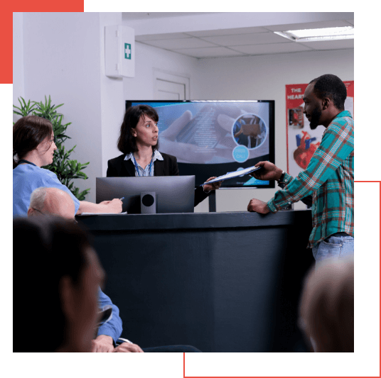  Three people are at a reception desk; a woman behind the desk appears to be assisting a man and another woman in front. In the background, there are pamphlets, medical posters, and information on Managed Detection and Response (MDR) services.